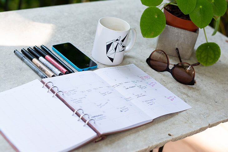 Auf einem Marmortisch liegt ein aufgeschlagener Kalender mit einigen Einträgen. Darüber liegen verschiedenfarbige Stifte aufgereiht, ein Smartphone, eine Kaffeetasse und eine Sonnenbrille. Daneben steht eine Topfpflanze.