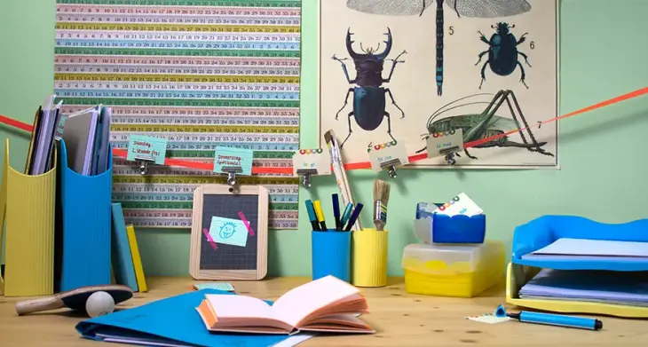 Auf einem Kinderschreibtisch stehen ein gelber und blauer Stehsammler, in denen Hefte sind, ein blauer und ein gleber Stifteköcher mit Stiften und Pinsel, gelbe und blaue Karteiboxen und Ablagen der Marke HAN. Auf dem Tisch liegt eine blaue Kladde und ein aufgeschlagenes Buch. An der Wand hinter dem Schreibtisch hängt ein Poster mit Käfern und eins mit Zahlen.
