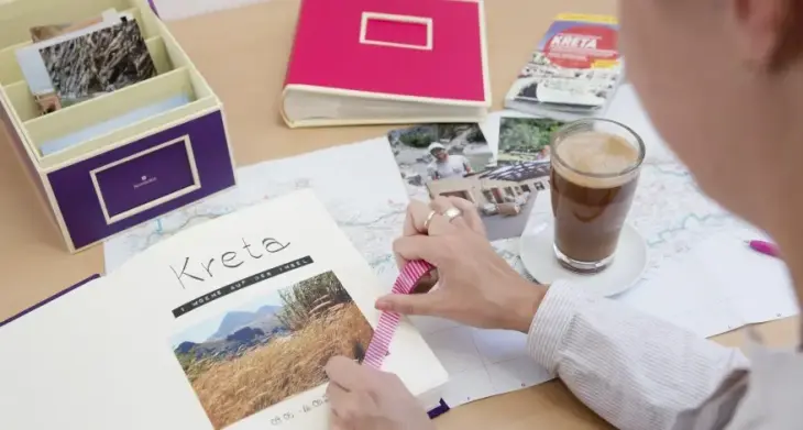 Auf einem Tisch liegt ein Fotoalbum, in das eine Frau Fotos aus einem Urlaub einklebt und mit Washi-Tape verziert. Außerdem liegen noch ein zugeschlagenes Fotoalbum und eine Fotobox der Marke Semikolon auf dem Tisch, sowie diverse Urlaubsfotos und eine Landkarte und ein Reiseführer.