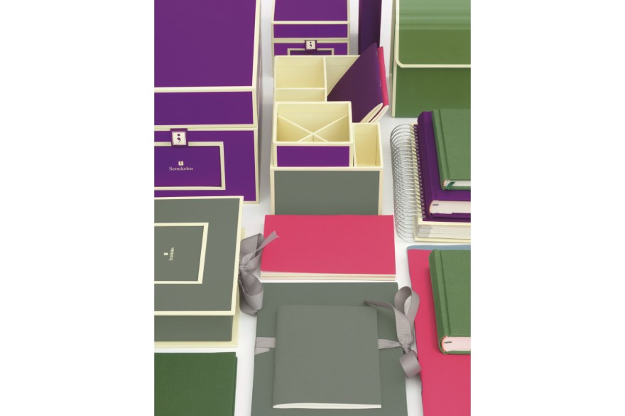 Diverse Produkte wie Aufbewahrungsboxen, Notizbücher,, Stifteköcher von Semikolon in pink, grün, grau und lila liegen auf einem Tisch ausgebreitet.