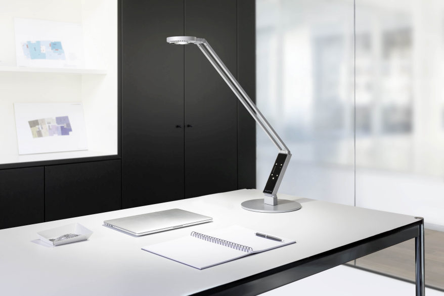 Auf einem Schreibtisch in einem Büro steht die Schreibtischlampe "Luctra" der Marke Durable. Auf dem Schreibtisch liegt ein zugeklapptes Notebook, ein geöffneter Spiralblock und einen Stiftschale.