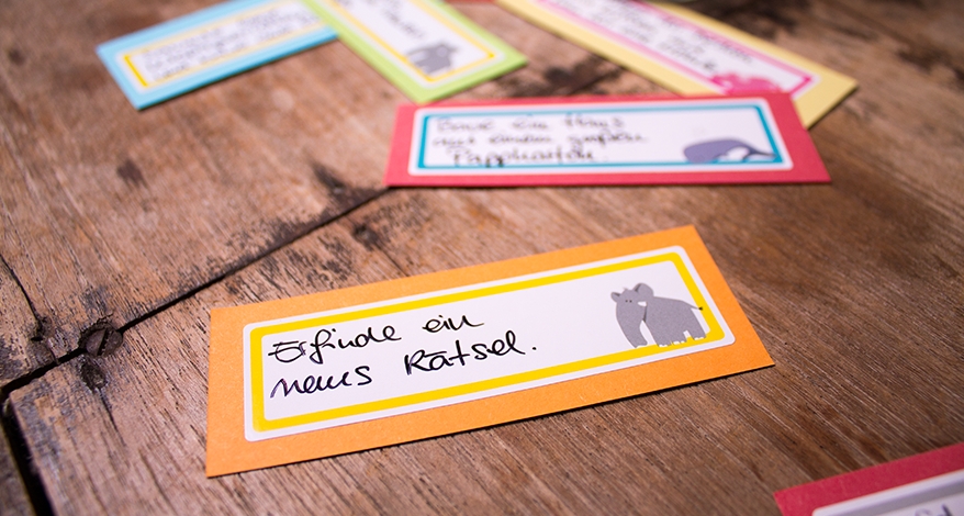 Nahaufnahme eines Aufgaben-Kärtchens zur Beschäftigung von Kindern. Auf dem Sticker, der auf das Kärtchen geklebt ist, ist ein Elefant zu sehen und es steht als Aufgabe geschrieben: "Erfinde ein neues Rätsel."
