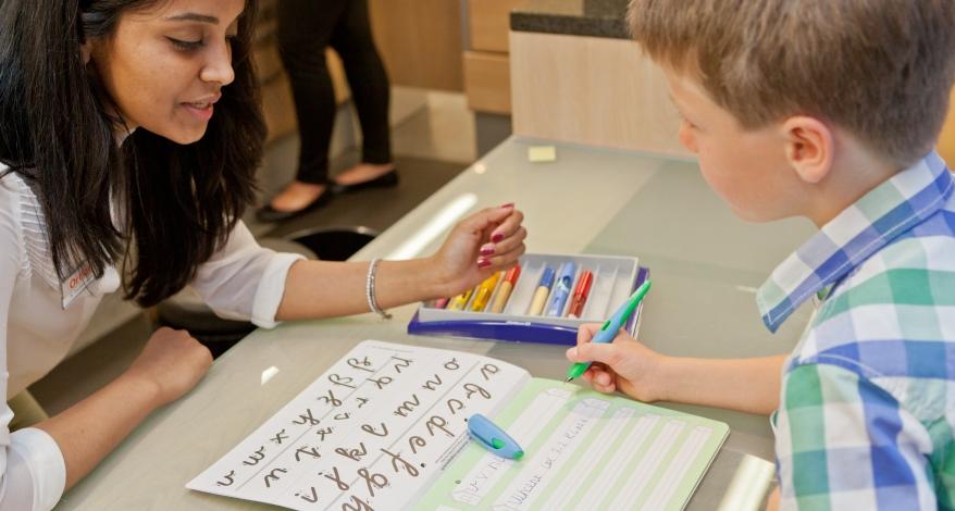 Ein Junge sitzt in einem Schreibwarengeschäft an einem Tisch und probiert mit Schreibübungen einen Füller aus. Eine Verkäuferin sitzt ihm gegenüber und schaut ihm dabei zu.