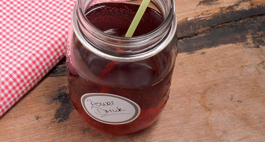 Auf einem dunklen Holztisch steht ein Weckglas, welches mit einem Getränk gefüllt und einem runden Avery Zweckform Küchenetikett beklebt ist. Darauf steht "Power Drink". Daneben liegt ein rot-weiß kariertes Küchentuch.