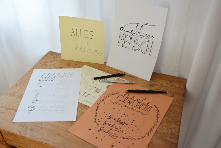 Auf einem kleinen Holztisch liegen mehrere verschiedenfarbige Papiere, auf denen Sprüche und Buchstaben gelettert wurden. Es liegen außerdem verschiedene Stifte auf den Papieren.