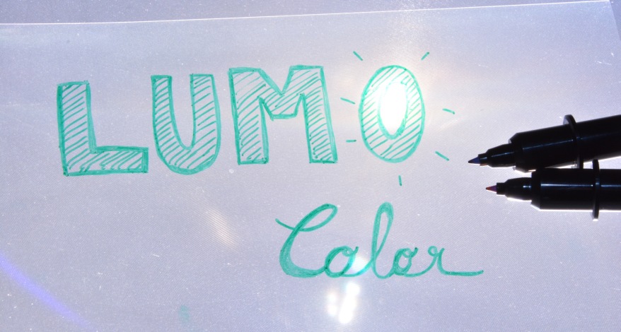 Auf einer Overhead-Folie ist mit grünem Lumocolor Faserschreiber der Marke Staedtler das Wort "Lumocolor" gelettert. Die geöffneten Spitzen von 2 Faserschreibern sind daneben zu sehen.