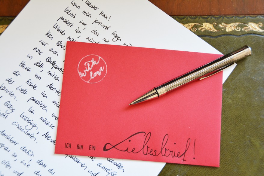 Auf einer alten Schreibunterlage liegt ein hangeschriebener Brief. Darauf liegt ein roter Umschlag, auf den gelettert steht "Ich bin ein Liebesbrief" und ein Stempel "With love" gestempelt wurde. Auf dem Brief liegt ein Kugelschreiber der Marke Parker.