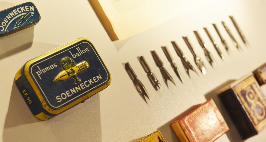 Auf einem Tisch liegen diverse Schreibfedern der Marke Soennecken. Daneben liegt die Metallschachtel mit der Aufschrift "Soennecken", in der die Federn aufbewahrt werden. Außerdem liegen weitere kleine Schachteln auf dem Tisch.