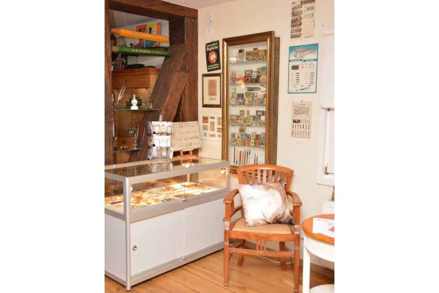 Ecke im Museum für Schreibkultur mit einer Vitrine mit Verpackungen von Stahlfedern, einer Vitrine mit Stahlfedern, einem Holzstuhl und diversen Bildern.