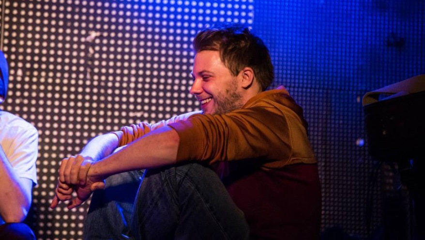 Der Poetry Slammer Christian Ritter sitzt auf einer Bühne. Er sitzt im Schneider sitzt und lacht. Er sitzt seitlich zur Kamera.