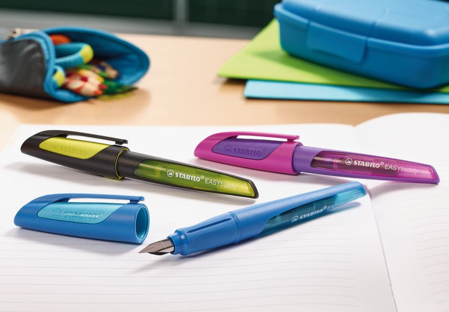 Drei bunte Stabilo EASYbuddy-Füller auf einem Notizheft, umgeben von Schulutensilien wie Federmappe und Brotdose.