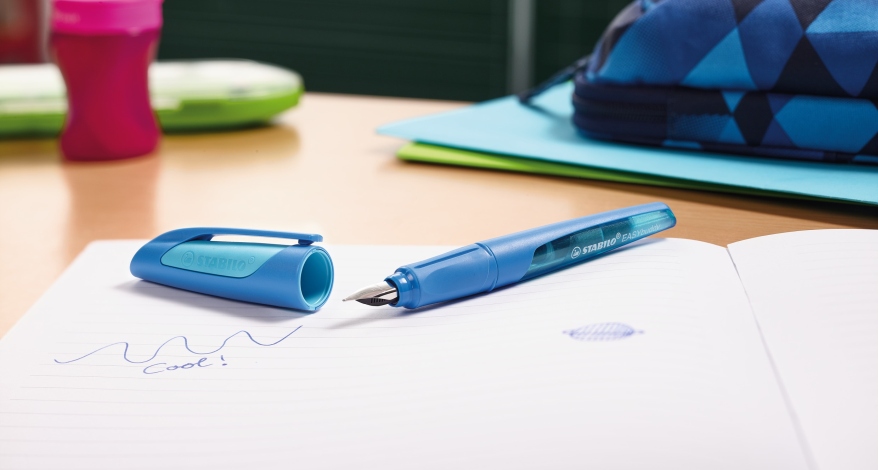 Ein blauer Stabilo-Füller ohne Kappe  leigt auf einem Notizheft. Darin ist mit Tinte eine geschwungene Linie zusehen und das Wort 'Cool!