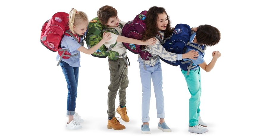4 Grundschulkinder stehen in einer Reihe seitlich nebeneinander und halten sich an den Schultern fest. Jedes Kind trägt einen ergobag Schulranzen auf dem Rücken.