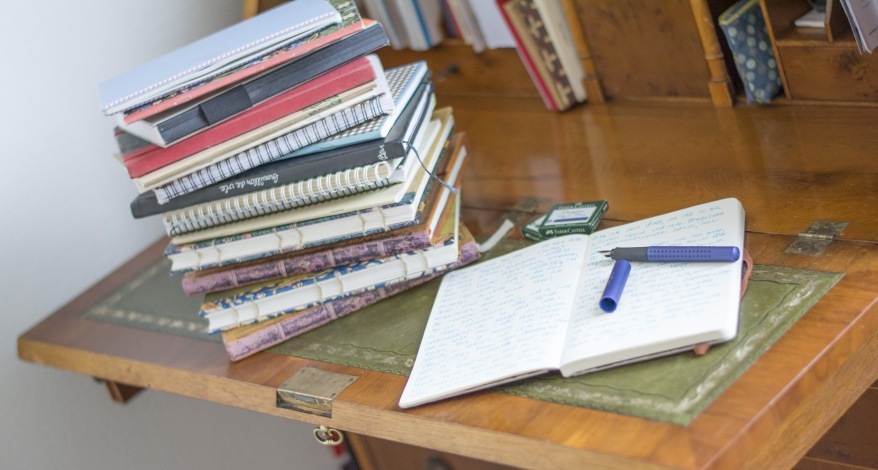 Auf einem Sekretär liegen ein Stapel Notizbücher und ein aufgeschlagenes Tagebuch. Auf der Seite des Tagebuchs liegt ein offener Füller.