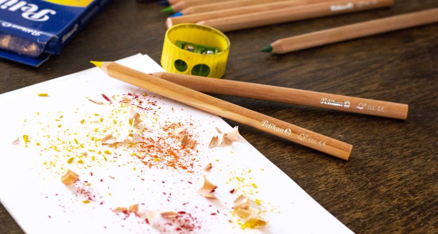 Auf einem Holztisch liegen ein Blatt Papier mit Spitzerresten, Buntstiften der Marke Pelikan und einem Spitzer.