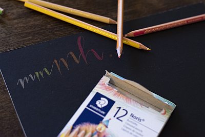 Buntes „mmmh“ auf schwarzem Papier, daneben Buntstifte und Verpackung von Staedtler