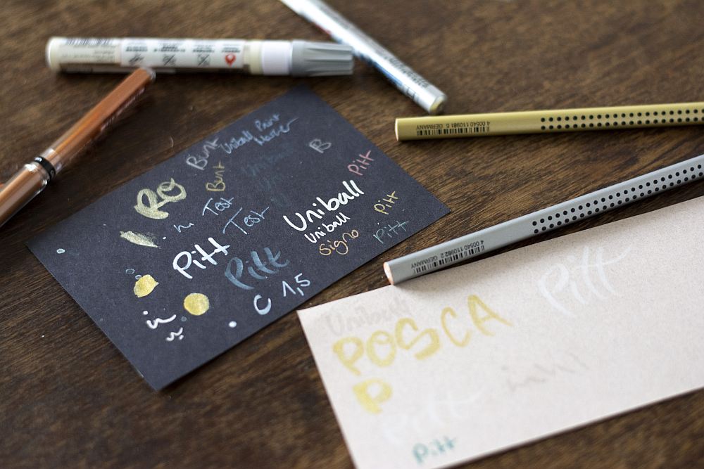 Testblätter mit verschiedenen Stiften auf dunklem und hellem Papier, umgeben von Stiften von Faber-Castell.