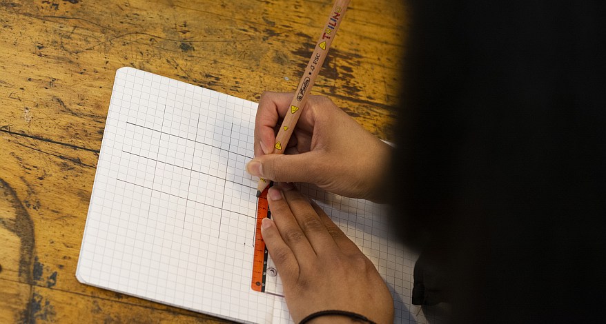 Eine Person hält einen Trilino-Buntstift der Marke Herlitz in der rechten Hand und malt mit Hilfe eines Lineals ein Gitternetz auf ein Blatt kariertes Papier. 