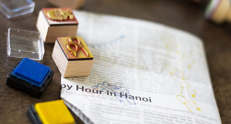 Auf einem dunklen Holztisch liegt einer zu einer Tüte gefalteten Zeitung. Diese wurde mit Maskingtape und Stempeln verziert. Daneben liegen geöffnete Stempelkissen und ein benutzter Stempel.