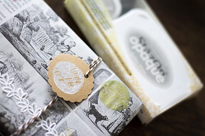Ein Close-up eines Geschenks, welches mit Zeitungspapier verpackt und mit Washitape und Stempeln verziert wurde. Mit gold-weißem Bäckergarn ist ein Geschenkanhänger aus Kraftpapier am Geschenk befestigt. Dieser wurde mit einem weißen Stempelmotiv bestempelt. Im Hintergrund ist unscharf eine Verpackung mit einem weißen Stempelkissen von StazOn zu sehen. 