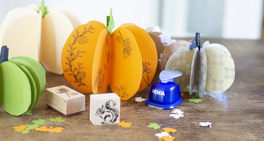Gebastelte Papierkürbisse in verschiedenen Farben und mit Stempeln verziert. Davor stehen diverse Stempel und Stanzer der Marke Heyda.