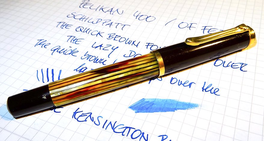 Ein schwarzer Füller mit goldenen Streifen der Marke Pelikan liegt auf einem karierten Papier, auf das mit blauer Tinte ein Text geschrieben wurde. Neben dem Füller prangt ein Tintenkleks.
