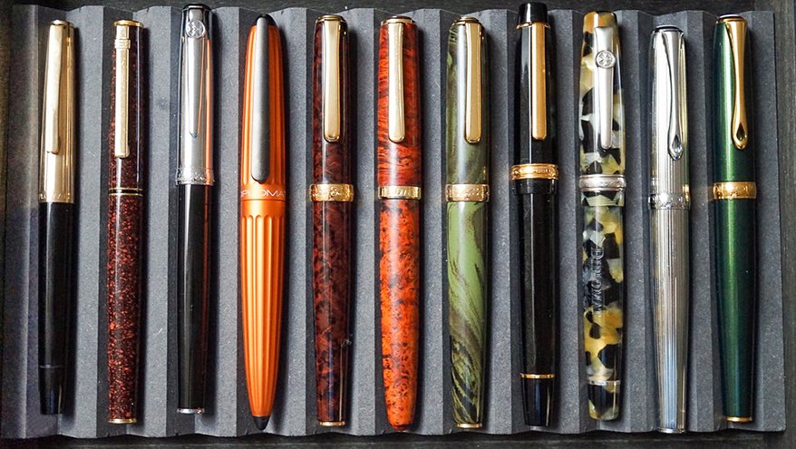 11 Füller liegen in einem grauen, mit Stoff ausgekleideteten, Etui.  Die Füller haben die unterschiedlichsten Farben, Formen und Verzierungen. 