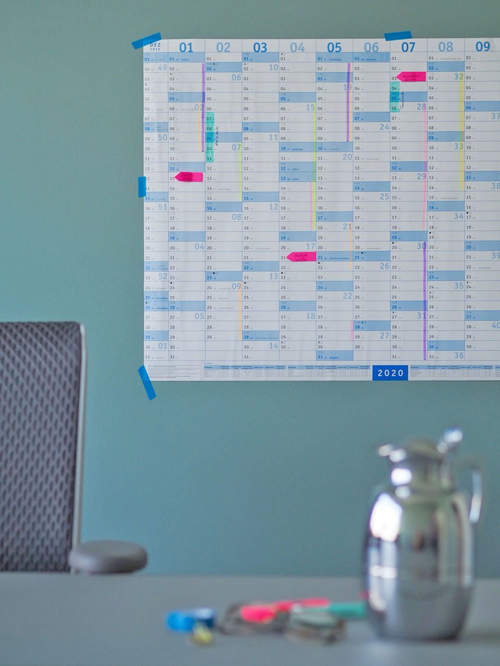 Auf einer blauen Wand ist ein Wandkalender der Marke Athesia angebracht. Dieser wurde mit verschiedenfarbigen Markern und Tapes beklebt. Im Vordergrund ist ein Bürostuhl und eine Kaffeekanne zu erkennen. Auf einem Schreibtisch liegen außerdem verschiedene Tapes und Stifte.