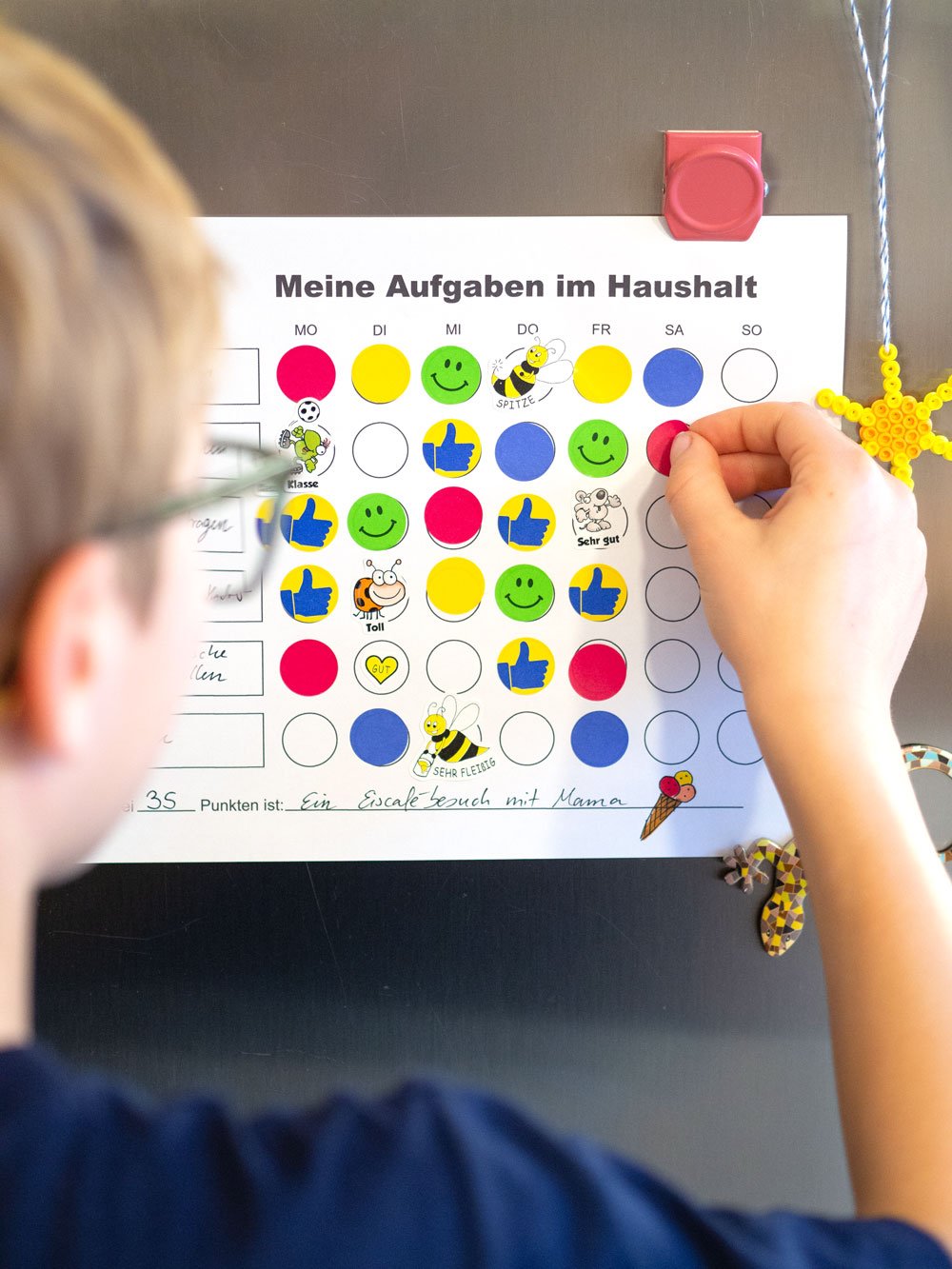 Ein Junge klebt einen roten Sticker auf den Haushaltsplan für eine erledigte Aufgabe. Drumherum kleben gelbe Punkte, grüne Smileys, Bienen-Sticker u.v.m. Überschrift auf dem Plan: Meine Aufgaben im Haushalt.