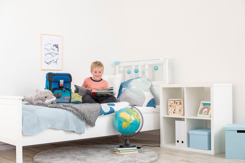 Ein Junge im Grundschulalter sitzt in seeinem Kinderzimmer auf seinem Bett und blättert in einem Buch. Neben ihm steht sein Step by Step Schulranzen. Vor seinem Bett steht ein Globus.