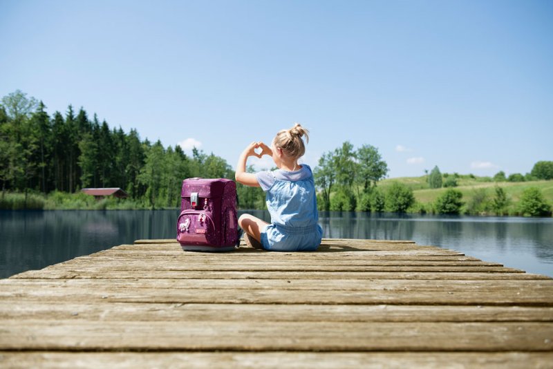 Ein Mädchen sitzen auf einem Steg vor einem See. Neben ihr steht ein Step by Step Schulranezen. Das Mädchen bildet mit ihren Händen ein Herz.