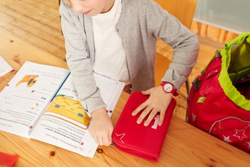 Ein Kind sitzt an einem Tisch. Vor ihm liegt ein aufgeschlagenes Schulheft und das Kind öffnet gerade ein Federmäppschen der Marke ergobag. Neben ihm steht ein ergobag Schulranzen.