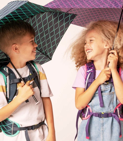Ein Junge und ein Mädchen im Grundschul-Alterstehen nebeneinander und lachen sich an. Beide halten einen Schirm der Marke ergobag in der Hand.