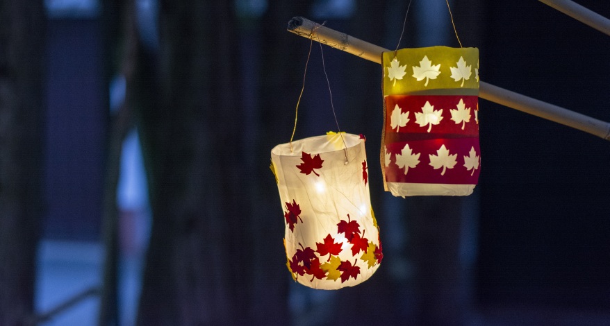 Zwei leuchtende Laternen mit bunten Ahornblättern an einem Ast in der Dunkelheit.