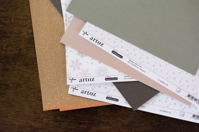 Verschiedenfarbige Papiere mit weihnachtlich glitzernder Anmutung der Marke Artoz liegen auf einem Holztisch.