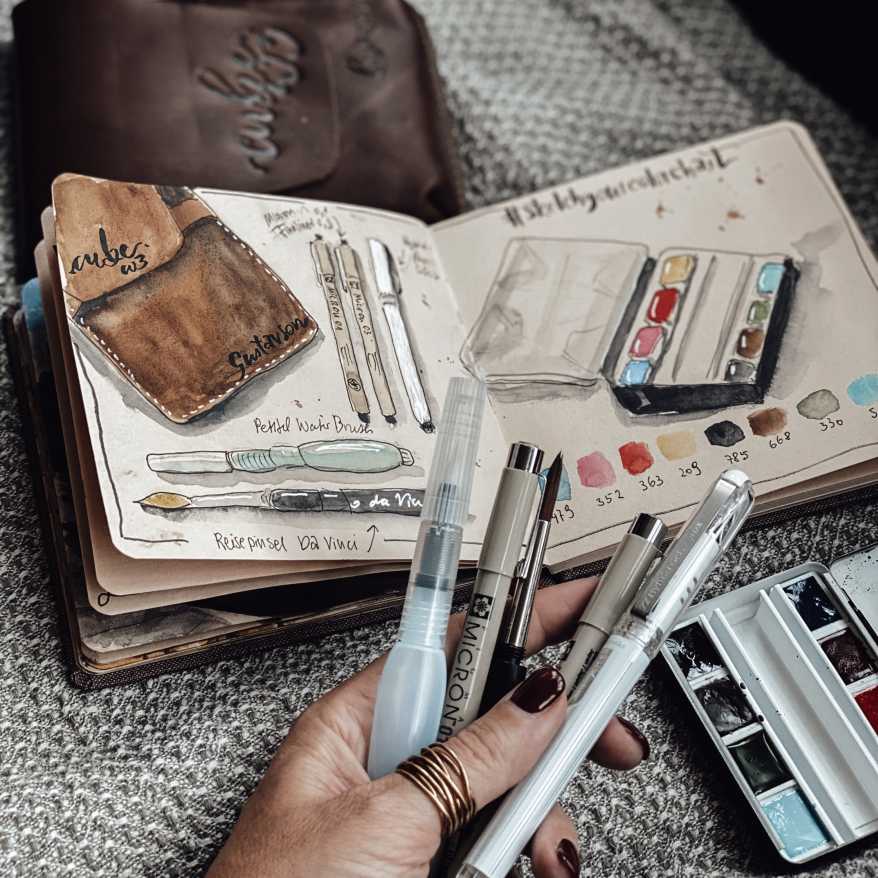 In einem Skizzenbuch sind die Seiten mit Aquarelmotiven bemalt. Eine Hand hält diverse Stifte und Pinsel fest.
