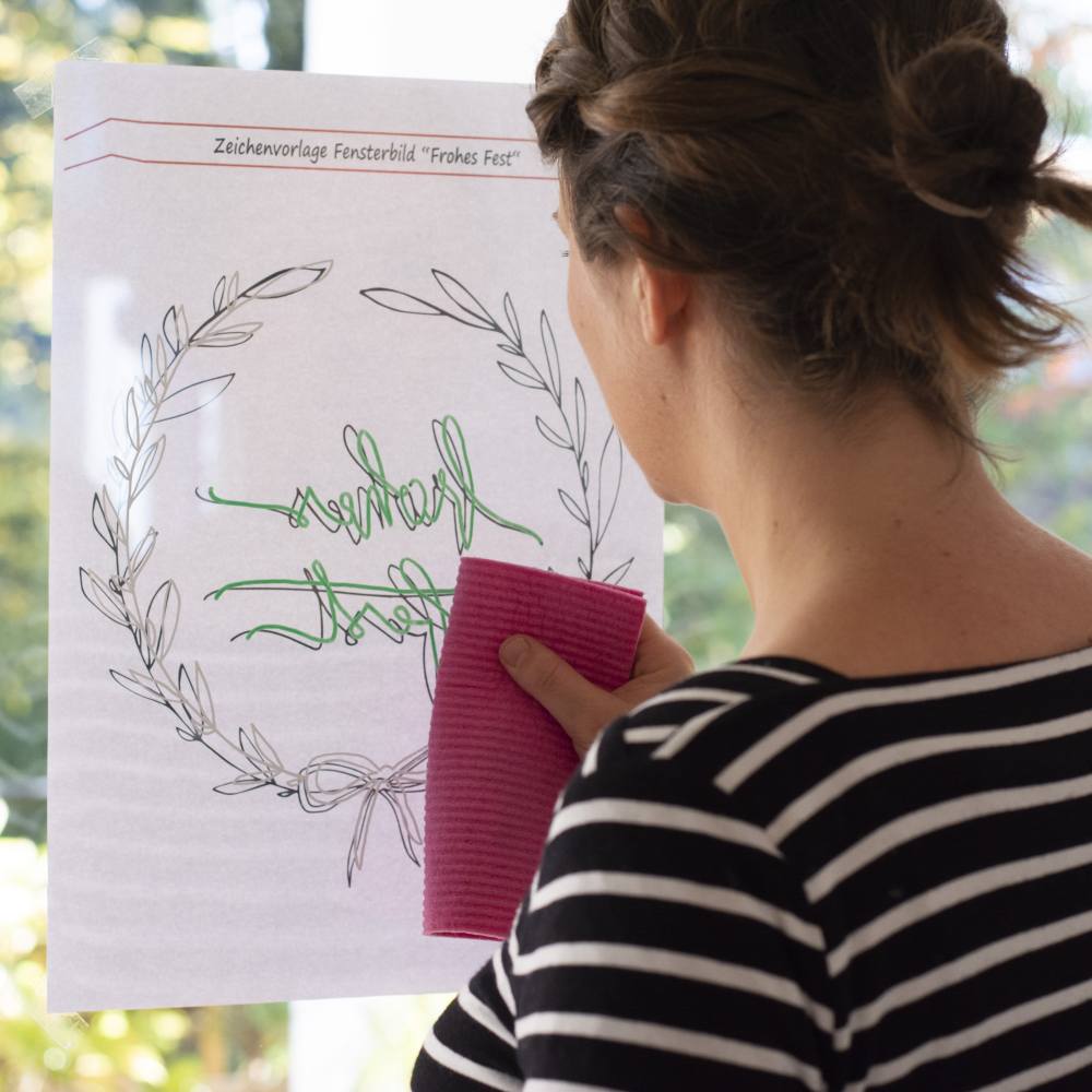 Eine junge Frau hat die Malvorlage für das weihnachtliche Fensterbild von außen ans Fenster geklebt die Konturen nachgemalt. Nun wischt sie sie mit einem Tuch wieder weg.
