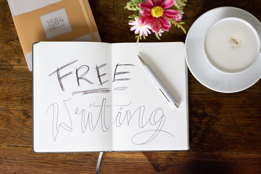Flatlay von aufgeschlagenem Notizbuch, in dem "Free Writing" als Handlettering steht. Umgeben von Kaffeetasse und Blumenstrauß.