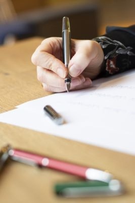 Nahaufnahme von Hand, die schreibt. Gezeigt wird der Dreifingergriff mit Füllhalter Pura von Pelikan, im Vordergrund liegen weitere Schreibgeräte und Stifte .