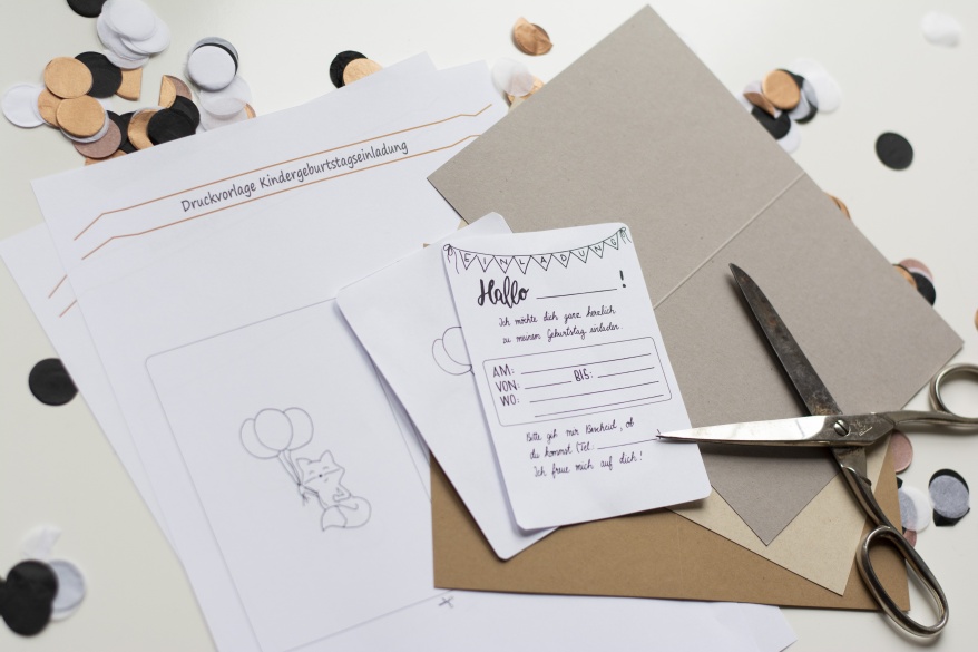 Einladungskarte zum Kindergeburtstag. Ausgeschnittene Druckvorlage mit Einladungstext, Schere und Klappkarte liegen auf Konfetti