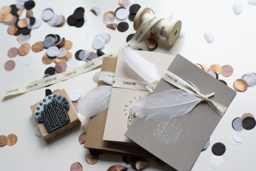 Einladungskarte zum Kindergeburtstag: Klappkarte mit Dekoband und Kunstfeder liegt auf Konfetti. Stempel mit Geburtstagstorte liegt daneben.