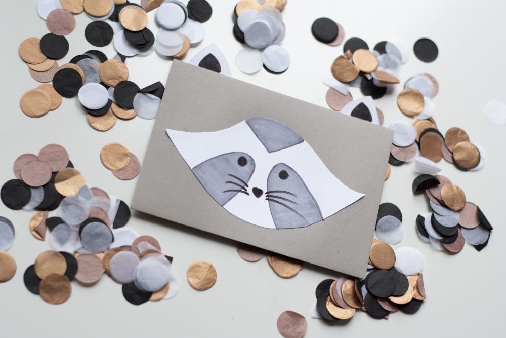 Einladungskarte zum Kindergeburtstag: Selbst gebastelte Do it yourself-Karte als Waschbär liegt auf Konfetti