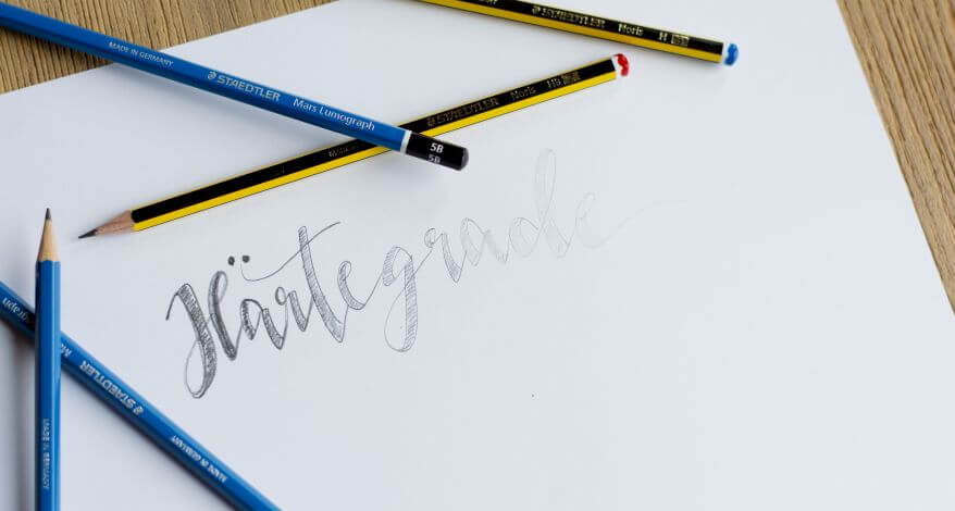 Auf einem Blatt Papier steht in Handlettering das Wort "Härtegrade", die Schrift verläuft von dunkel nach hell. Drumherum liegen die Bleistifte Noris und Lumograph der Marke Staedtler.