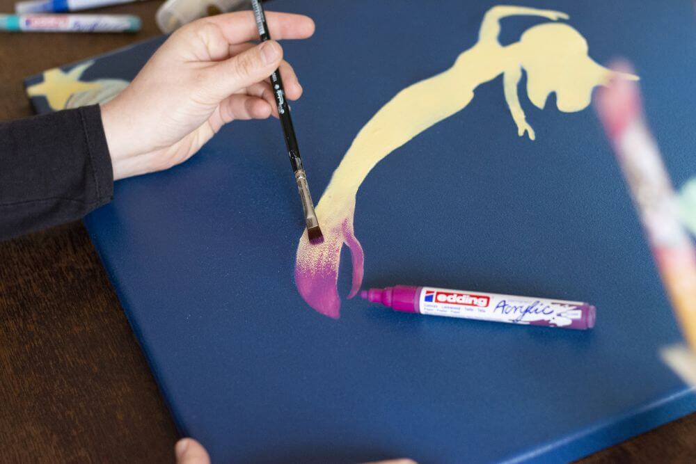 Eine Hand malt den Fischschwanz einer Meerjungfrau mit Acrylfarben von Edding auf Leinwand.