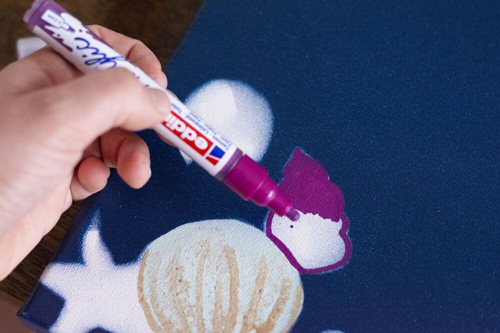 Nahaufnahme einer Hand, die mit einem Acrylmarker von Edding eine Seeschnecke auf eine Leinwand malt.