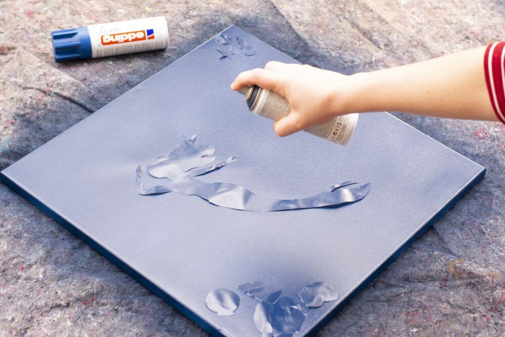 Eine Hand sprüht blaue Sprühfarbe von Edding auf eine Leinwand, auf der eine Schablone aus Papier in Form einer Meerjungfrau liegt.