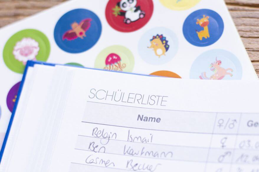Auf einem Tisch liegt ein Stickerbogen mit Tiermotiven. Darauf liegt ein Lehrerkalender mit einer ausgeüllten Schülerliste.