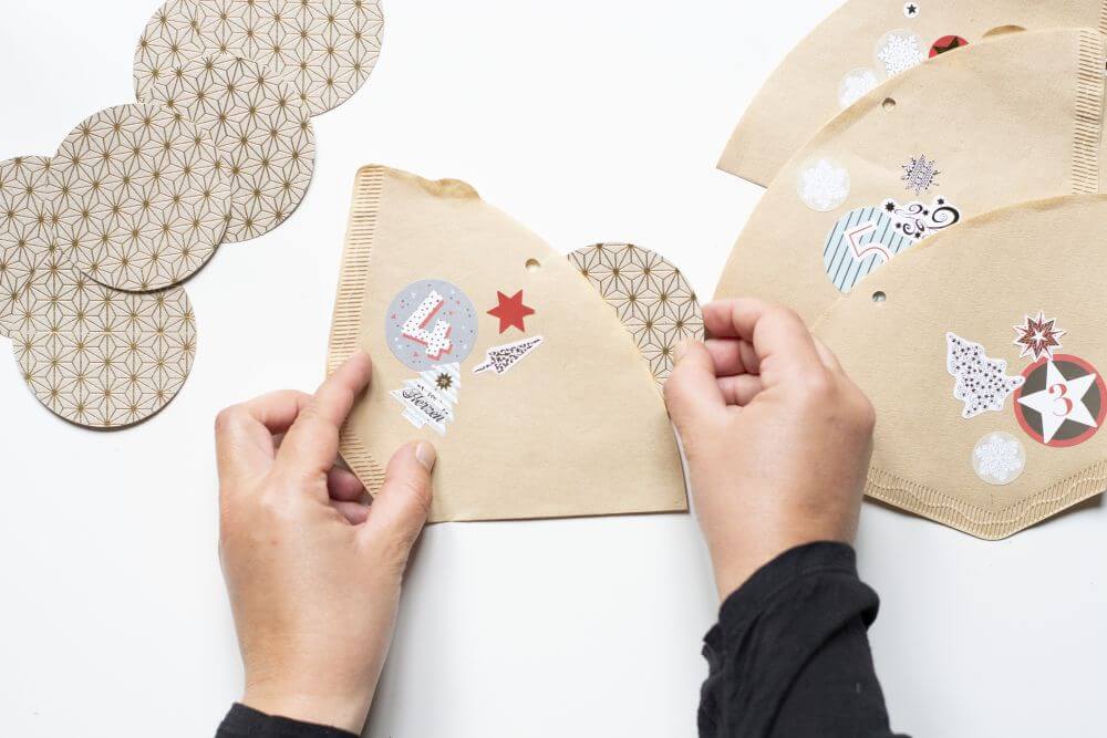 Ein kreisförmige Motivkarton, der mit einem Spruch beschriftet ist, wird in einen Kaffeefilter gesteckt. Auf diesem kleben Sticker mit Weihnachtsmotiven sowie die Zahl vier. 