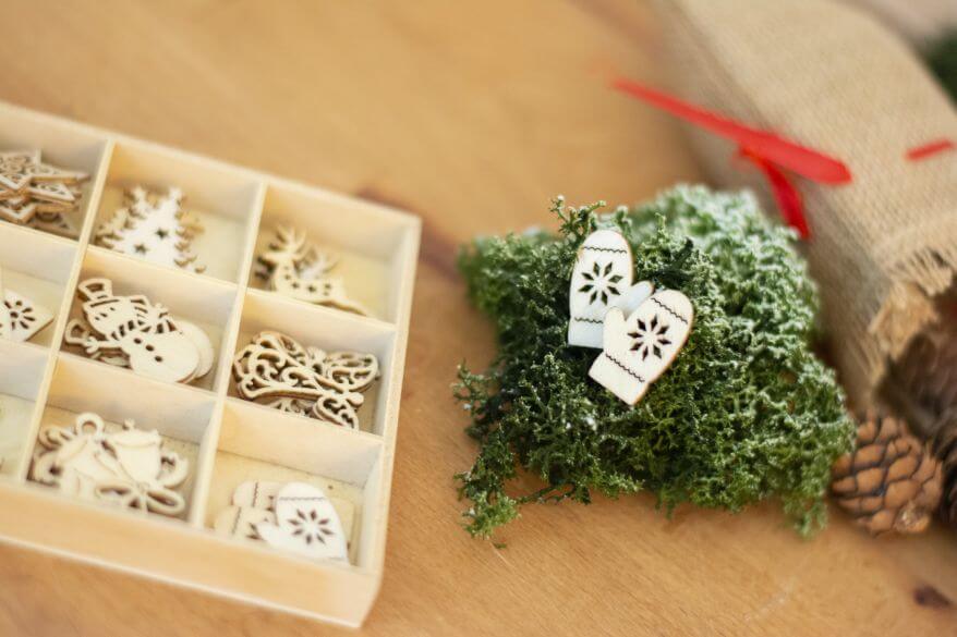 Auf Parkettboden liegend sieht man Dekomaterialien, mit denen eine Wichteltür zur Weihnachtszeit ergänzt wird: eine Box mit kleinen Holzornamenten in weihnachtlichen Motiven (z.B. Handschuhe) von Heyda liegt neben etwas Islandmoos.