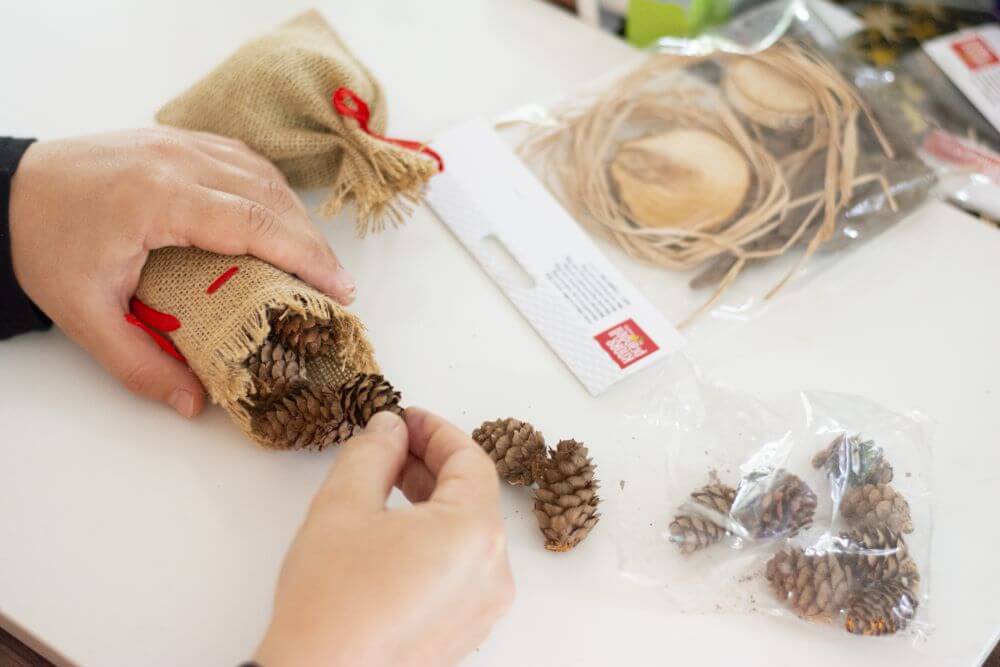 Blick auf einen Schreibtisch: Eine Hand hält ein kleines Jutesäckchen fest, die andere Hand befüllt es mit Lärchenzapfen zum Basteln von Knorr Prandell.
